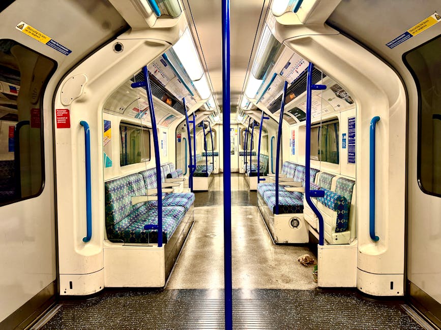 Interior view of a train carriage parked at Abbey Wood station, featuring clean, patterned fabric seats arranged in rows along both sides and facing each other. The carriage has a smooth, polished flo