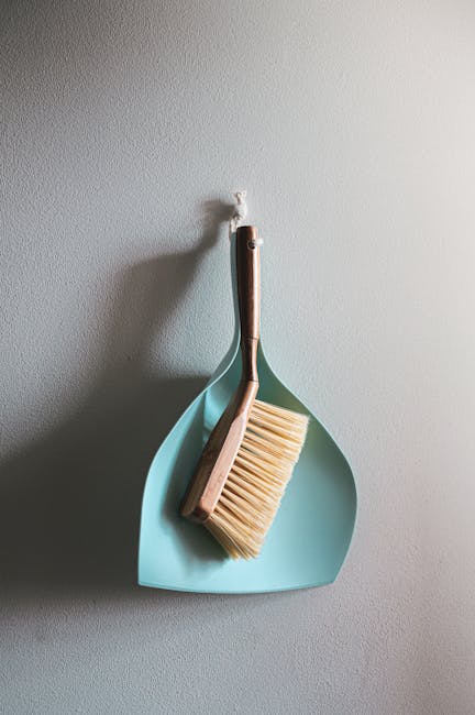 A person wearing a blue disposable glove is cleaning a remote control with a light blue cloth, focusing on the surface of the device. The remote is black, sitting on a flat, white surface, possibly a