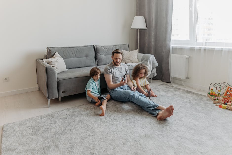 A woman with curly hair dressed in casual clothing is engaged in surface cleaning using a vacuum cleaner with a long hose attachment. She is in a living room with a wooden floor and a patterned area r