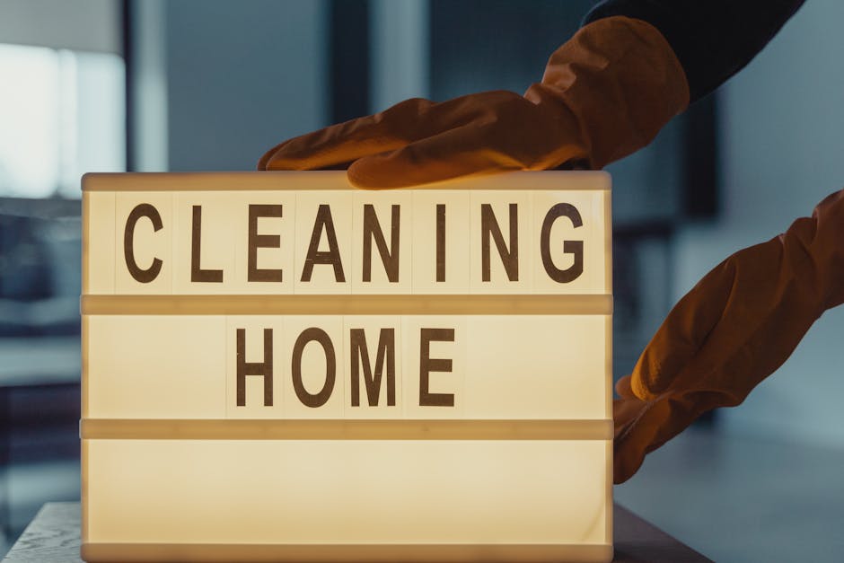 A person wearing brown cleaning gloves is placing or adjusting a lit-up signboard with the words 'CLEANING HOME' on a wooden table inside a modern living room. The background features a sofa, a coffee